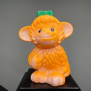 Puede incluir: Un juguete de peluche de mono naranja con un lazo verde en la cabeza. El mono tiene una cara sonriente y ojos grandes.
