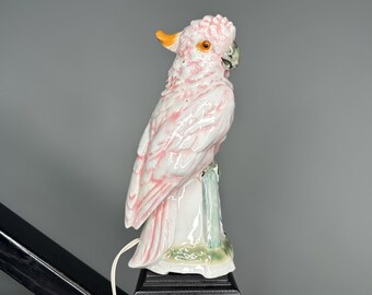May include: A ceramic parrot figurine in shades of pink, white, and orange. The bird is perched on a decorative base with a light cord extending from the bottom. The parrot has detailed feathers and a realistic appearance.
