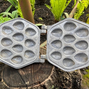 Op de afbeelding: Een vintage metalen wafeltang met 18 individuele mallen in de vorm van amandelen. De tang is open en staat op een boomstronk.
