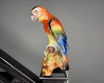 May include: A colorful ceramic parrot sculpture perched on a decorative base. The parrot features vibrant orange, yellow, green, and blue plumage. The sculpture is a decorative item, likely for display.