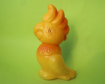 May include: A yellow plastic toy cockatoo with a red crest and a flower design on its head. The toy is standing on a green surface.