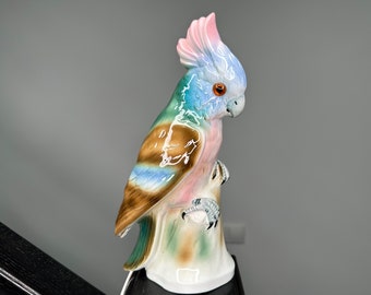 May include: A colorful porcelain figurine of a cockatoo perched on a branch. The bird is painted in shades of pink, blue, green, and brown.