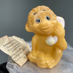 Puede incluir: Un mono de juguete de goma amarillo con un corazón blanco en el pecho. El mono está sentado con la mano derecha en la barbilla y la mano izquierda sosteniendo un objeto blanco. El juguete está etiquetado como "Коопера Э" y tiene el código de producto "2-Д-2-234".