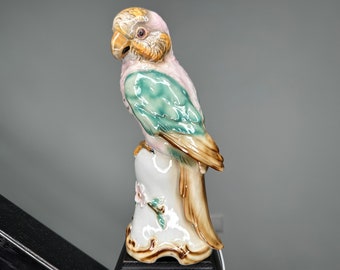 May include: A ceramic parrot figurine with green, pink, and brown accents. The parrot is perched on a white base with floral details.