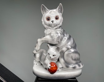 May include: A white ceramic figurine of a mother cat and her kitten. The mother cat is sitting with her paw on the kitten's back. The kitten is playing with a red ball.