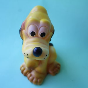 Puede incluir: Un perro de juguete de goma amarillo con ojos negros y nariz negra. El perro está sentado sobre una superficie azul claro.