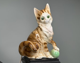 May include: A ceramic cat ornament with brown and tan stripes, green eyes, and a white chest. The cat is seated, holding a small green ball with its paw. The figurine has a glossy finish. The background is grey.