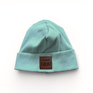 Könnte beinhalten: Eine helltürkisfarbene Beanie-Mütze mit gerippter Struktur. Ein braunes quadratisches Patch mit den Worten "ciao KAKAO" ist auf der Vorderseite aufgenäht. Die Mütze ist aus weichem Material und für den Alltag geeignet.