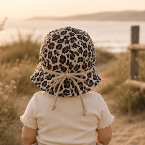 Könnte beinhalten: Sonnenhut mit Leopardenmuster und beiger Schleife und Bindebändern. Der Hut wird von einem Kind getragen, mit einem cremefarbenen Hemd. Der Hintergrund zeigt eine Strandszene mit Holzzaun.
