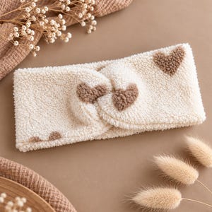 Könnte beinhalten: Ein cremefarbenes Stirnband aus Sherpa-Fleece mit gedrehtem Design auf der Vorderseite und einem wiederkehrenden Muster aus kleinen, flauschigen braunen Herzen. Der weiche, strukturierte Stoff sorgt für eine plüschige Optik mit gleichmäßig verteilten Herzmotiven.
