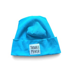 Könnte beinhalten: Eine leuchtend türkisfarbene Rippstrickmütze mit umgeschlagenem Saum. Ein weißes rechteckiges Patch mit den Worten "TROUBLE MAKER" in schwarzen Blockbuchstaben ist auf der Vorderseite aufgenäht. Die Mütze ist einfarbig und vor weißem Hintergrund.
