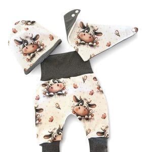 Könnte beinhalten: Ein dreiteiliges Baby-Set mit Mütze, Lätzchen und Hose. Das Set ist cremefarben mit einem sich wiederholenden Muster aus Cartoon-Kühen und Blumen. Die Bündchen und der Bund sind dunkelgrau.