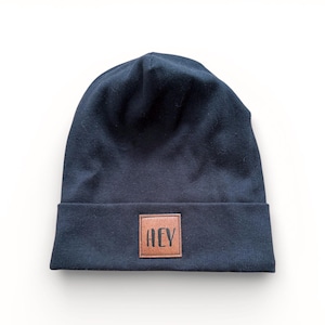 Könnte beinhalten: Marineblaue Beanie-Mütze mit einem braunen Lederaufnäher mit der Aufschrift "HEY". Die Mütze ist aus einem weichen Strickstoff gefertigt und hat einen umgeschlagenen Saum. Die Mütze befindet sich auf weißem Hintergrund.