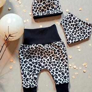 Könnte beinhalten: Ein Baby-Kleidungsset mit Leopardenmuster. Das Set besteht aus einer Hose, einer Mütze und einem Halstuch. Die Hose hat einen schwarzen Bund und Bündchen. Mütze und Halstuch sind ebenfalls mit Leopardenmuster bedruckt.