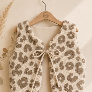 Könnte beinhalten: Eine ärmellose Weste aus cremefarbenem Sherpa-Fleece mit braunem Leopardenmuster. Die Weste verfügt über einen Frontverschluss mit zwei langen, passenden cremefarbenen Stoffbändern, die in der Mitte der Brust zu einer Schleife gebunden sind.