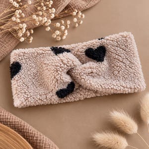 Könnte beinhalten: Ein beiges Stirnband aus Sherpa-Fleece mit gedrehtem Vorderteil und einem wiederkehrenden Muster aus kleinen schwarzen Herzen. Der strukturierte Stoff hat eine weiche, flauschige Optik und das Accessoire ist mit einem Knoten detail versehen.