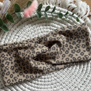 Könnte beinhalten: Ein braunes und beigefarbenes Stirnband mit Leopardenmuster. Der Stoff ist gerippt und vorne verdreht. Das Stirnband liegt auf einem weißen, gewebten Platzset mit dekorativem Grün und einem rosa, flauschigen Akzent.
