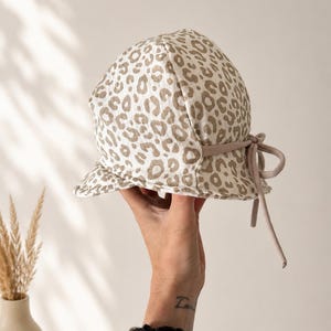 Könnte beinhalten: Sonnenhut mit Leopardenmuster in Beige und Weiß. Er hat eine runde Krone, eine kleine Krempe und eine hellbraune Kordel. Der Hut wird von einer Hand gehalten.