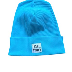 Könnte beinhalten: Eine leuchtend türkisfarbene Rippstrick-Beanie-Mütze. Die Mütze hat einen umgeschlagenen Saum und einen kleinen weißen rechteckigen Aufnäher mit dem schwarzen Aufdruck "TROUBLE MAKER".