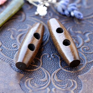 Könnte beinhalten: Zwei braune Holz-Toggle-Knöpfe mit jeweils zwei Löchern. Die Knöpfe befinden sich auf einer braunen Lederoberfläche mit einem dekorativen Muster.
