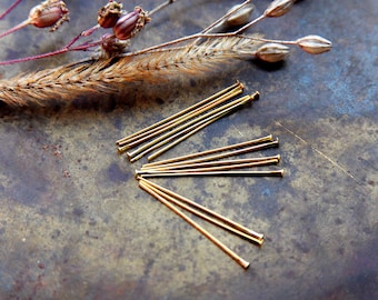 May include: A collection of gold-colored head pins arranged on a textured, dark metal surface. The pins are straight with a small, rounded head. Dried seed pods and a sprig of dried grass are in the background, suggesting a craft or jewelry-making theme.