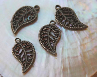 Könnte beinhalten: Vier bronzefarbene Blattanhänger mit filigranen Details liegen auf einer weißen, schillernden Oberfläche. Eine rosa Blume ist ebenfalls zu sehen. Die Anhänger haben eine Schlaufe zum Aufhängen, ideal für Schmuckherstellung oder Bastelarbeiten.