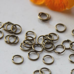 Könnte beinhalten: Eine große Menge kleiner, goldfarbener Sprungringe. Die Ringe sind alle gleich groß und auf einer weißen Oberfläche in einem zufälligen Muster angeordnet.