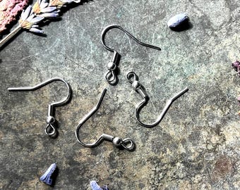 May include: Close-up of several silver-colored earring hooks, scattered on a textured, gray surface. The hooks have a simple, curved design with a small ball and loop at the bottom. Dried lavender and small blue flower buds are also present.