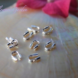 Könnte beinhalten: Nahaufnahme von silberfarbenen Quetschperlen auf einer weißen, schillernden Oberfläche. Die Perlen sind klein und haben an einem Ende eine Schlaufe.