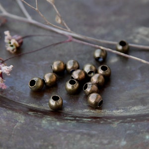 May include: A close-up of a group of small, round, brass beads scattered on a dark brown surface. The beads have a slightly aged, antique look.