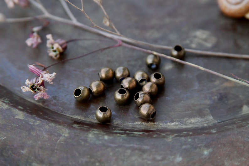 May include: A close-up of a group of small, round, brass beads scattered on a dark brown surface. The beads have a slightly aged, antique look.