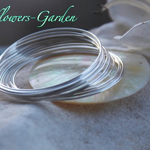 Könnte beinhalten: Eine Ansammlung silberfarbener Drahtarmbänder, gestapelt und auf einer perlmuttartigen Oberfläche angeordnet. Die Armbänder sind dünn und glänzend und reflektieren das Licht. Der Text "Venusflowers-Garden" befindet sich oben links.