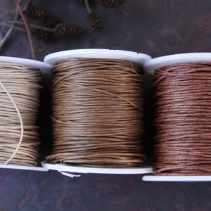 Könnte beinhalten: Drei Spulen gewachster Schnur in verschiedenen Brauntönen. Die Spulen sind aus weißem Kunststoff und die Schnur ist fest gewickelt.