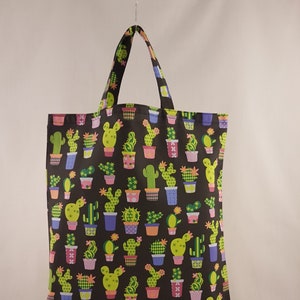 Op de afbeelding: Een zwarte draagtas met een herhaald patroon van verschillende cactussen in kleurrijke potten. De cactussen zijn groen en geel en de potten zijn roze, blauw en oranje. De tas heeft een bijpassende handgreep.