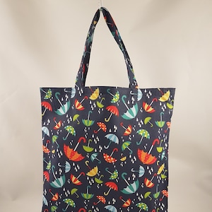 Puede incluir: Una bolsa de mano azul marino con estampado de paraguas de colores. Los paraguas son rojos, amarillos, verdes y azules con lunares blancos. La bolsa tiene dos asas y un fondo blanco con gotas de lluvia blancas.