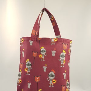 Può includere: Una borsa tote color borgogna con un motivo ripetuto di cavalieri, castelli e bandiere dei cartoni animati. I cavalieri indossano armature d'argento e tengono spade e scudi.