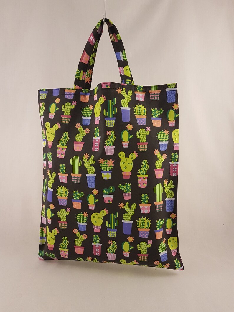 Op de afbeelding: Een zwarte draagtas met een herhaald patroon van cactussen in kleurrijke potten. De cactussen zijn groen en geel, de potten blauw, paars en oranje. De tas heeft een bijpassende handgreep en is gemaakt van stevige stof.