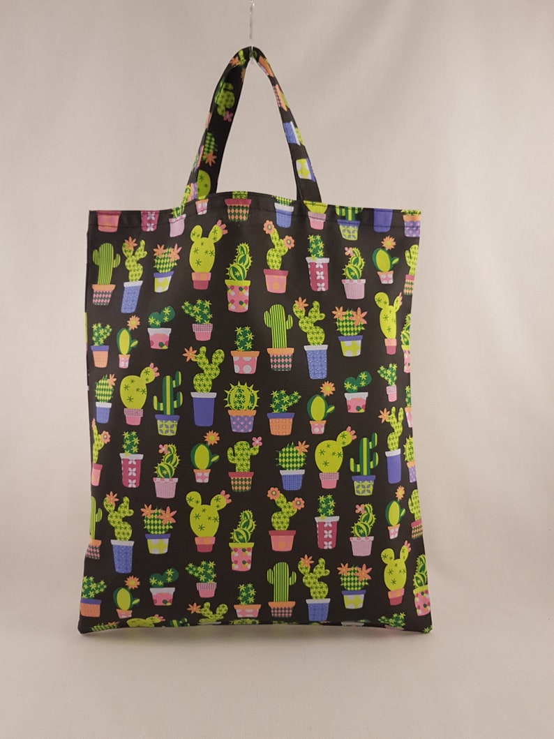 Op de afbeelding: Een zwarte draagtas met een herhaald patroon van verschillende cactussen in kleurrijke potten. De cactussen zijn groen en geel en de potten zijn roze, blauw en oranje. De tas heeft een bijpassende handgreep.