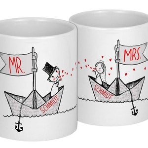 Pu&ograve; includere: Due tazze in ceramica bianca con illustrazioni in nero e rosso di una coppia in barche di carta. Il testo "MR." e "MRS." &egrave; sulle bandiere delle barche. Il testo "SCHMIDT" &egrave; sul fondo di ogni barca. La coppia si sta mandando cuori.