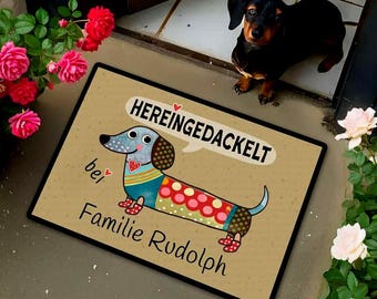 May include: A rectangular doormat with a cartoon dachshund design. The dachshund is blue, red, and yellow, with the text "HEREINGEDACKELT" in a speech bubble. The text "bei Familie Rudolph" is below the dog.