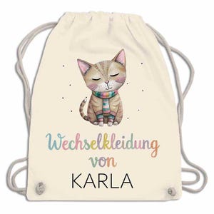 May include: A cream-colored drawstring backpack featuring a watercolor illustration of a cat wearing a striped scarf. The text "Wechselkleidung von KARLA" is printed in colorful letters below the cat. The bag has gray drawstrings.