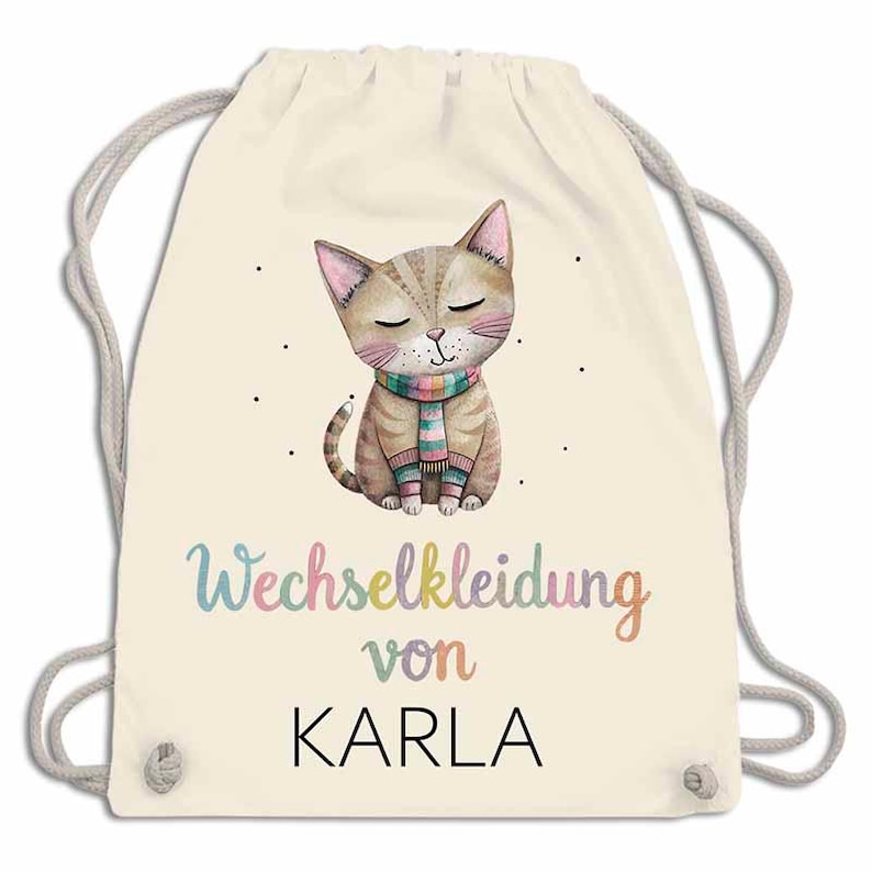 May include: A cream-colored drawstring backpack featuring a watercolor illustration of a cat wearing a striped scarf. The text "Wechselkleidung von KARLA" is printed in colorful letters below the cat. The bag has gray drawstrings.