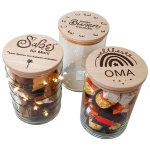 May include: Three glass jars with wooden lids. One jar contains cookies and fairy lights, another is empty, and the third holds wrapped candies. The lids are engraved with German text and designs.