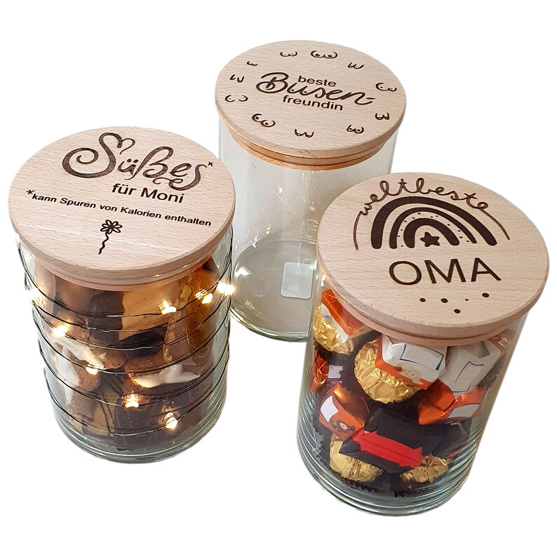 May include: Three glass jars with wooden lids. One jar contains cookies and fairy lights, another is empty, and the third holds wrapped candies. The lids are engraved with German text and designs.