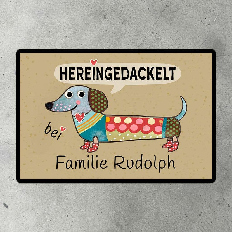 Eine personalisierte Fußmatte mit einem Dackel-Motiv und dem Spruch 'Hereingedackelt' auf dem Boden vor einer Haustür.
