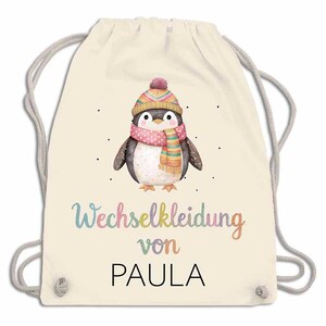 May include: A cream-colored drawstring backpack featuring a watercolor penguin wearing a pink scarf and a colorful hat. The text "Wechselkleidung von PAULA" is written in a rainbow font.