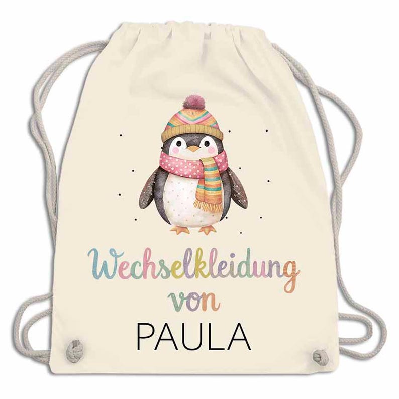 May include: A cream-colored drawstring backpack featuring a watercolor penguin wearing a pink scarf and a colorful hat. The text "Wechselkleidung von PAULA" is written in a rainbow font.