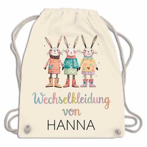 May include: A cream-colored drawstring backpack featuring a whimsical illustration of three anthropomorphic bunnies in colorful outfits and boots. The text "Wechselkleidung von HANNA" is printed below the bunnies in a playful font.