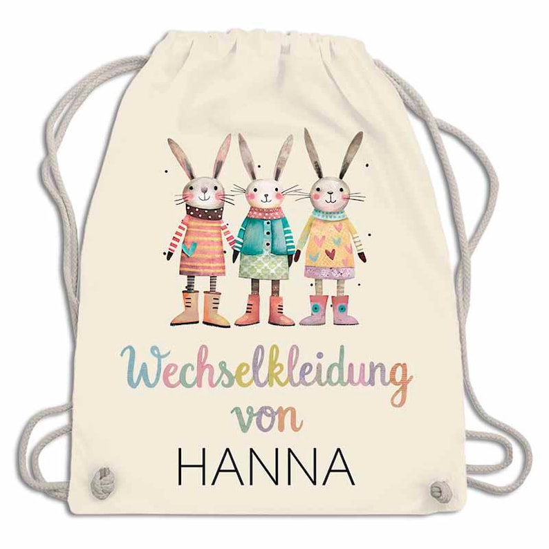 May include: A cream-colored drawstring backpack featuring a whimsical illustration of three anthropomorphic bunnies in colorful outfits and boots. The text "Wechselkleidung von HANNA" is printed below the bunnies in a playful font.