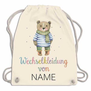 May include: A beige drawstring backpack featuring a cartoon bear wearing a striped shirt, scarf, and pants. The text "Wechselkleidung von NAME" is written in colorful script below the bear. The bag has gray drawstrings.